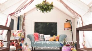 白いグランピングテントの内観。三角の天井からグリーンを飾ったシャンデリアが下がり、中央にはくすみブルーのヴィンテージソファとカラフルなクッション、足元にはエスニック柄のラグが敷かれた、華やかでボヘミアンな空間。「モビリティリゾートもてぎ完全ガイド｜料金・アクセス・グランピング」の文字入り。栃木県茂木町のリゾート施設（旧ツインリンクもてぎ）を紹介するおでかけガイド記事のアイキャッチ画像。