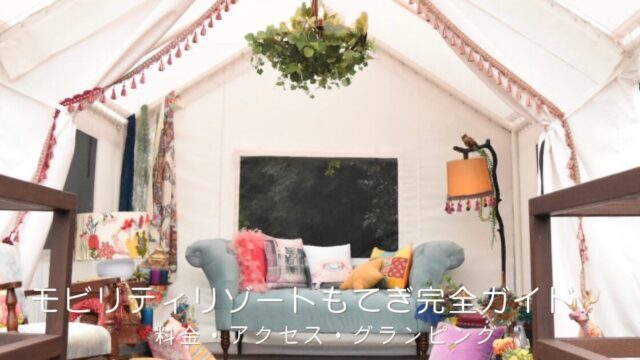 白いグランピングテントの内観。三角の天井からグリーンを飾ったシャンデリアが下がり、中央にはくすみブルーのヴィンテージソファとカラフルなクッション、足元にはエスニック柄のラグが敷かれた、華やかでボヘミアンな空間。「モビリティリゾートもてぎ完全ガイド｜料金・アクセス・グランピング」の文字入り。栃木県茂木町のリゾート施設（旧ツインリンクもてぎ）を紹介するおでかけガイド記事のアイキャッチ画像。